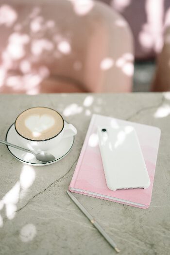 A table with a cup of coffee with a heart shaped cream dollop in the center, a pink ombre notebook and cell phone