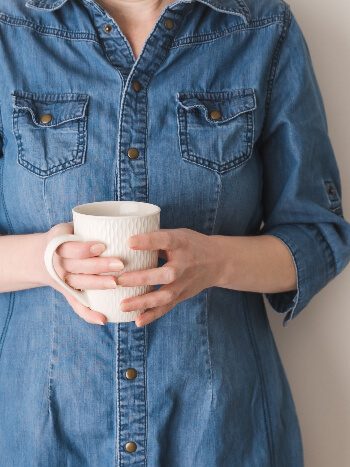A woman in a blue denim shirt leaning against a wall holding a coffee mug
