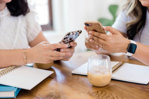 two women sitting at a table with their notebooks open and searching on their cell phones